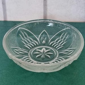 Puede incluir: Cuenco de vidrio transparente con exterior texturizado y diseño floral grabado en el interior. El cuenco tiene forma redonda y se apoya sobre una superficie verde. El diseño floral presenta una estrella central.