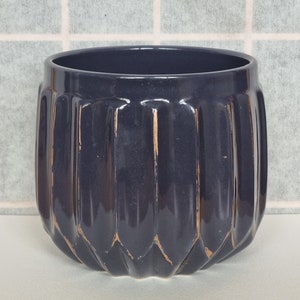 Puede incluir: Una maceta de cerámica azul oscuro con un diseño acanalado y detalles dorados. La maceta cilíndrica tiene una abertura ancha y una superficie texturizada. Esta maceta decorativa es adecuada para uso en interiores o exteriores.