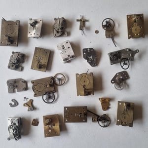 May include: A selection of antique watch components, including gears, plates, and balance wheels, in various shapes and sizes. The parts are made of brass and silver-coloured metal, arranged on a white surface. These vintage watch parts are suitable for restoration or crafting.