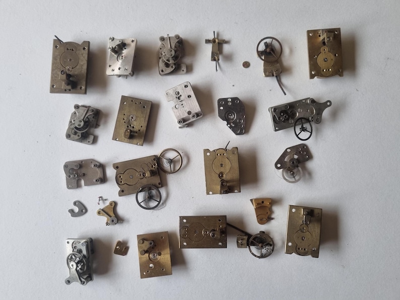 May include: A collection of vintage watch parts, including gears, plates, and balance wheels, in various shapes and sizes. The components are made of brass and silver-colored metal, arranged on a white surface. These antique watch components are ideal for restoration or crafting.