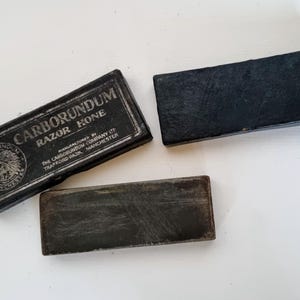 May include: Three vintage Carborundum Razor Hone sharpening stones. One is in its original black box with white text and a logo. The other two are rectangular, dark grey stones. The box reads "Carborundum Razor Hone" and "Manufactured by The Carborundum Company Ltd, Trafford Park, Manchester."