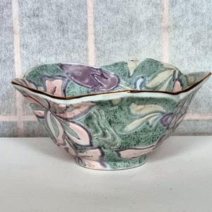 May include: A decorative ceramic bowl with a floral design. The bowl features a green, pink, and purple color palette with a gold rim. The floral pattern is embossed on the surface, adding texture to the piece.