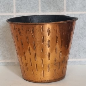 Puede incluir: Un macetero de cobre con textura martillada y un forro de plástico negro. El macetero es de estilo tradicional con una boca ancha y una base estrecha.