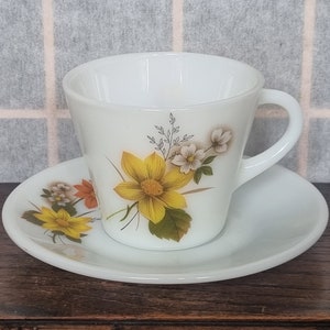 Puede incluir: Juego de taza y platillo blancos. La taza y el platillo están decorados con un diseño floral con flores amarillas y blancas con hojas verdes. La taza tiene un asa y se apoya en un platillo a juego.