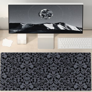 Op de afbeelding: Een zwart en grijs muismat met bloemenpatroon ligt op een licht houten bureau. De muismat is rechthoekig en heeft een gedetailleerd damastontwerp. Een wit toetsenbord en een computermonitor zijn ook zichtbaar.