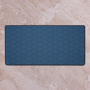 May include: A rectangular desk mat featuring a blue wave pattern with a black border. The wave design is a repeating pattern of semi-circles in a darker shade of blue. The mat is set against a light brown marble surface.