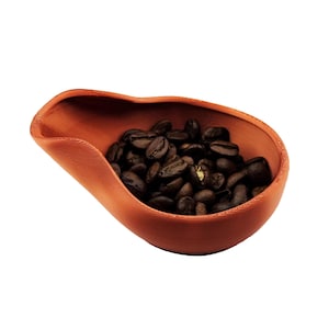 Puede incluir: Un cuenco en forma de cuchara, de color terracota, lleno de granos de café tostados oscuros. El cuenco tiene un diseño liso y curvado y está sobre un fondo blanco. Los granos de café son de color marrón oscuro.