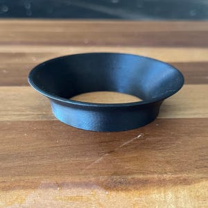 Puede incluir: Un objeto negro en forma de embudo circular, con una abertura ensanchada y una superficie texturizada. El objeto está hecho de un material sólido y descansa sobre una superficie de madera. El objeto parece ser una herramienta o un accesorio. El diámetro es de unos 7 cm.