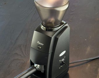 Puede incluir: Un molinillo de café negro Baratza Encore con una tolva oscura translúcida en la parte superior, un botón de encendido frontal y un dial lateral para los ajustes de molienda. La unidad incluye un mango de portafiltro negro en la zona de salida.