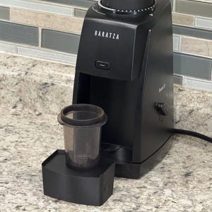 May include: A black Baratza Encore coffee grinder with a clear plastic grounds container. The grinder is on a countertop with a neutral-toned backsplash. The grinder has a dial and a power button.