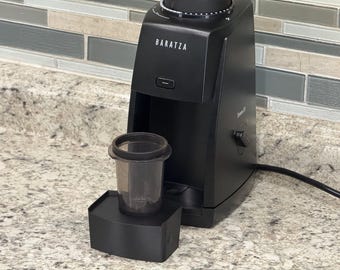 Puede incluir: Un molinillo de café negro Baratza Encore con un recipiente transparente de plástico para el café molido. El molinillo está sobre una encimera con un protector contra salpicaduras de tonos neutros. El molinillo tiene un dial y un botón de encendido.