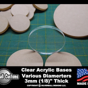 May include: Clear acrylic bases, 3mm (1/8") thick, in various diameters. The image shows a clear, circular acrylic base with several unfinished, round, cardboard bases on a green grid surface. The text "Clear Acrylic Bases" is visible.