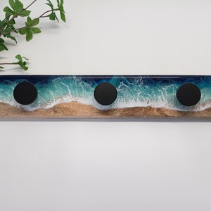 Könnte beinhalten: Ein Wand-Kleiderhaken aus Holz mit einem Harz-Ozeanwellen-Design. Der Haken hat drei schwarze runde Haken.