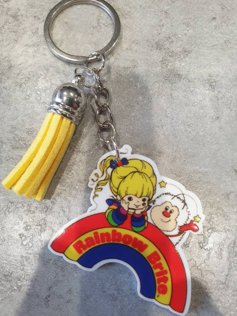 May include: A yellow tassel keychain with a rainbow and the words "Rainbow Brite" on it. The keychain features a cartoon image of Rainbow Brite and her friend, a white fluffy creature.