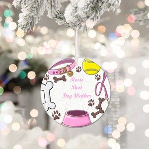 May include: A round Christmas ornament with a dog theme. The ornament features a pink dog collar, a yellow tennis ball, paw prints, and the text "Rosie Best Dog Walker". A pink dog bowl and a bone are also included.
