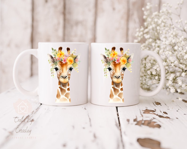 May include: Two white ceramic mugs with a watercolor illustration of a giraffe wearing a flower crown. The giraffe has a brown and tan spotted pattern. The mugs are on a white wooden surface.