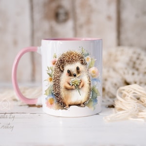 May include: A white ceramic mug with a pink handle and rim. The mug features a watercolor illustration of a hedgehog holding a flower. The background is a soft watercolor wash of blue and pink.