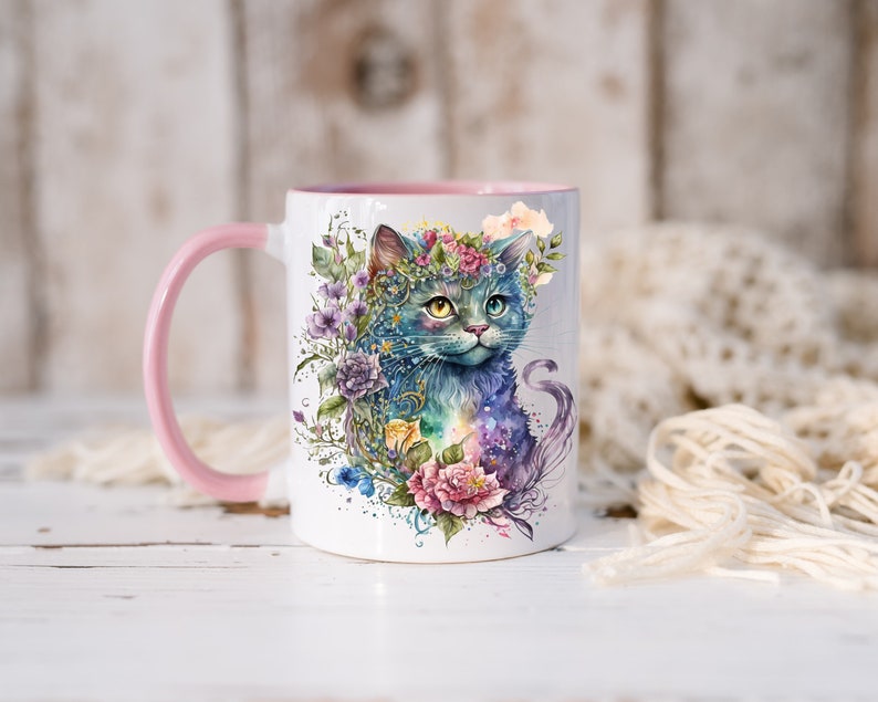 May include: A white ceramic mug with a pink rim and handle. The mug features a watercolor illustration of a blue cat with a flower crown, surrounded by colorful flowers and foliage.