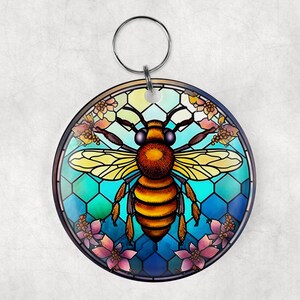 May include: A round keychain featuring a stained glass style bee with yellow and brown stripes, surrounded by blue and green honeycomb and pink flowers.