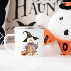 May include: White enamel mug featuring a cartoon duck dressed as a witch, holding a pumpkin and a staff. The duck wears a black witch hat with stars. The mug has a silver rim and a white handle. Halloween themed.