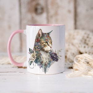 May include: A white ceramic mug with a pink handle and rim. The mug features a watercolor illustration of a tabby cat with flowers and foliage surrounding it.