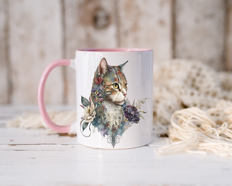 May include: A white ceramic mug with a pink handle and rim. The mug features a watercolor illustration of a tabby cat with flowers and foliage surrounding it.