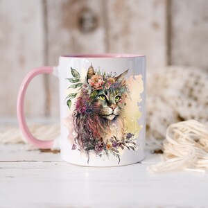 May include: A white ceramic mug with a pink rim and handle. The mug features a watercolor illustration of a cat wearing a flower crown. The cat has green eyes and brown fur. The background is a watercolor wash of yellow and pink.
