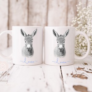 May include: Two white ceramic mugs with a black and white illustration of a donkey's head on each mug. The name "Laura" is written in blue cursive on the bottom of each mug.