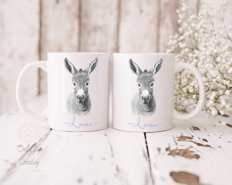 May include: Two white ceramic mugs with a black and white illustration of a donkey's head on each mug. The name "Laura" is written in blue cursive on the bottom of each mug.