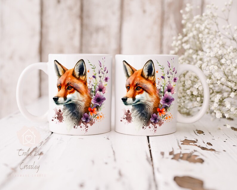 May include: Two white ceramic mugs with a watercolor design of a fox with purple and orange flowers. The mugs have a handle on the right side.