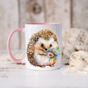 May include: A white ceramic mug with a pink rim and handle features a watercolor illustration of a hedgehog holding a bouquet of flowers. The hedgehog is brown with a light brown nose and black eyes.