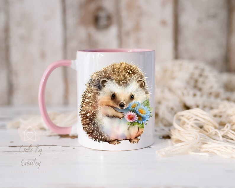 May include: A white ceramic mug with a pink rim and handle features a watercolor illustration of a hedgehog holding a bouquet of flowers. The hedgehog is brown with a light brown nose and black eyes.