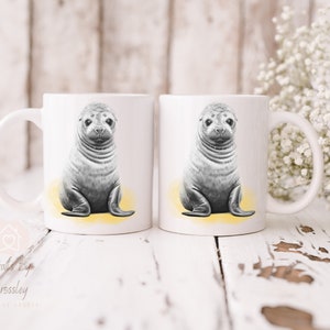 K&ouml;nnte beinhalten: Zwei wei&szlig;e Keramikbecher mit einer Schwarzwei&szlig;illustration eines Seehunds auf jedem Becher. Der Seehund sitzt auf einem gelben Hintergrund.
