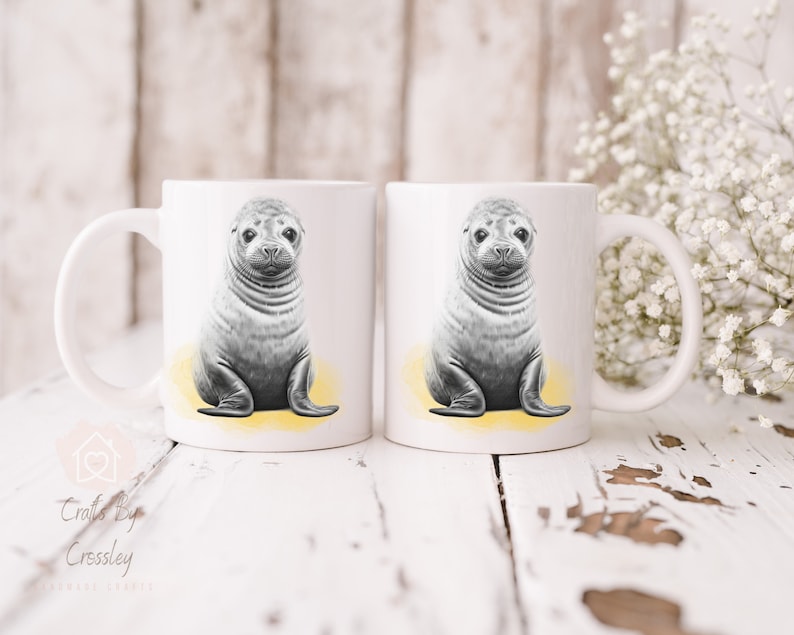 K&ouml;nnte beinhalten: Zwei wei&szlig;e Keramikbecher mit einer Schwarzwei&szlig;illustration eines Seehunds auf jedem Becher. Der Seehund sitzt auf einem gelben Hintergrund.