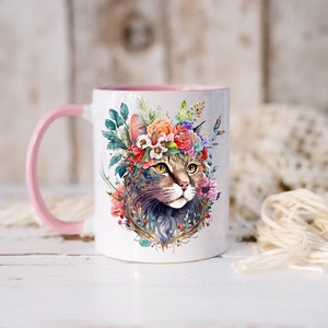 May include: White ceramic mug with a pink handle. The mug features a watercolor illustration of a cat wearing a floral crown. The cat has green eyes and a brown and gray tabby pattern.