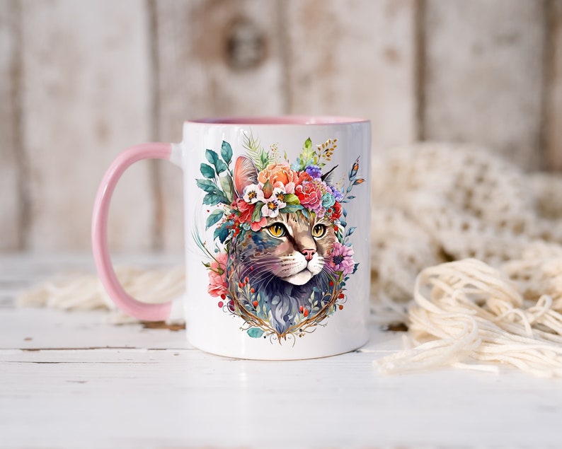 May include: White ceramic mug with a pink handle. The mug features a watercolor illustration of a cat wearing a floral crown. The cat has green eyes and a brown and gray tabby pattern.
