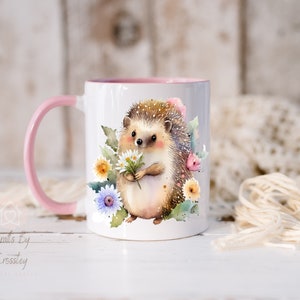 May include: A white ceramic mug with a pink rim and handle features a watercolor illustration of a hedgehog holding a bouquet of white daisies. The hedgehog is surrounded by pink, yellow, and blue flowers.