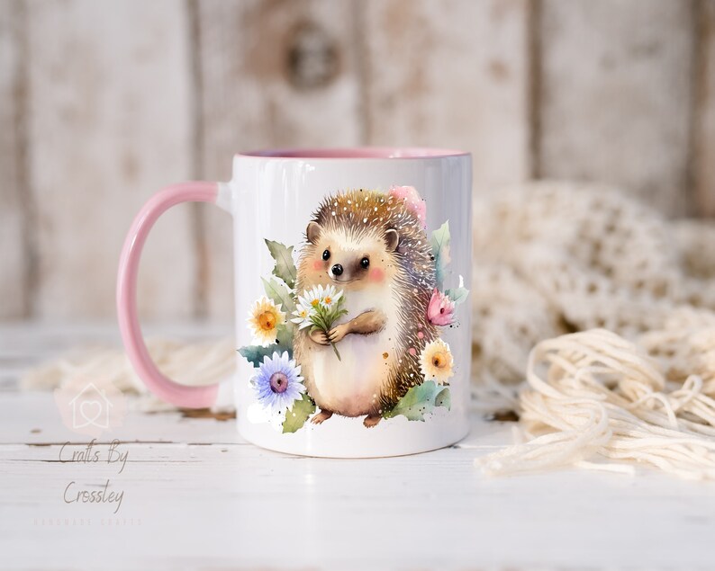 May include: A white ceramic mug with a pink rim and handle features a watercolor illustration of a hedgehog holding a bouquet of white daisies. The hedgehog is surrounded by pink, yellow, and blue flowers.
