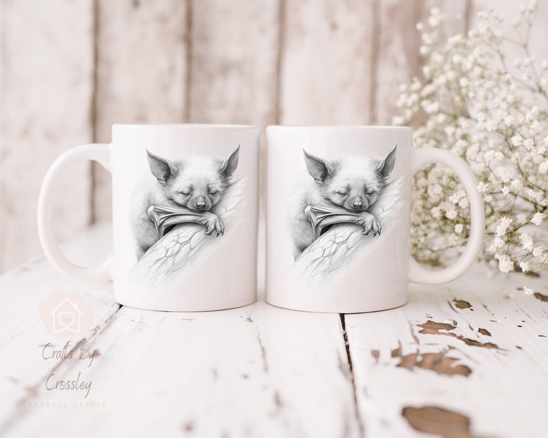 Puede incluir: Dos tazas de cer&aacute;mica blanca con una ilustraci&oacute;n en blanco y negro de un murci&eacute;lago durmiendo en cada taza. El murci&eacute;lago est&aacute; enroscado en una rama y tiene sus alas envueltas alrededor de s&iacute; mismo.