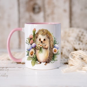 May include: A white ceramic mug with a pink rim and handle features a watercolor illustration of a hedgehog holding a bouquet of flowers. The hedgehog is brown with a white belly and has a pink nose. The flowers are white, pink, and blue.