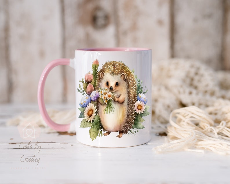 May include: A white ceramic mug with a pink rim and handle features a watercolor illustration of a hedgehog holding a bouquet of flowers. The hedgehog is brown with a white belly and has a pink nose. The flowers are white, pink, and blue.
