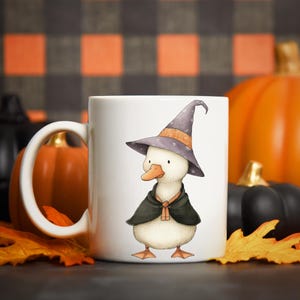May include: White ceramic mug featuring a whimsical illustration of a duck wearing a witch hat and cape. The duck is depicted with a light-colored body, orange feet and beak, and a purple hat. The mug is set against a backdrop of pumpkins and fall leaves.