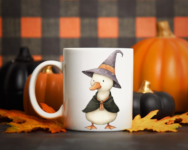 May include: White ceramic mug featuring a whimsical illustration of a duck wearing a witch hat and cape. The duck is depicted with a light-colored body, orange feet and beak, and a purple hat. The mug is set against a backdrop of pumpkins and fall leaves.