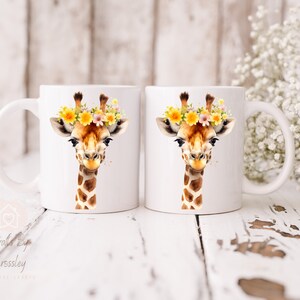 May include: Two white ceramic mugs with a watercolor illustration of a giraffe wearing a flower crown. The giraffe has brown spots and a light brown face. The mugs are on a white wooden surface.
