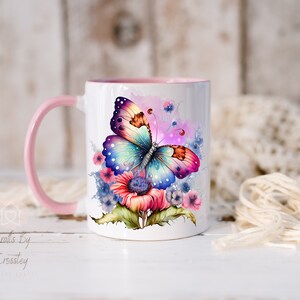 K&ouml;nnte beinhalten: Eine wei&szlig;e Keramiktasse mit einem rosa Henkel zeigt einen bunten Schmetterling mit schillernden Fl&uuml;geln und einem Blumenmuster. Der Schmetterling ist von rosa, blauen und gelben Blumen umgeben.