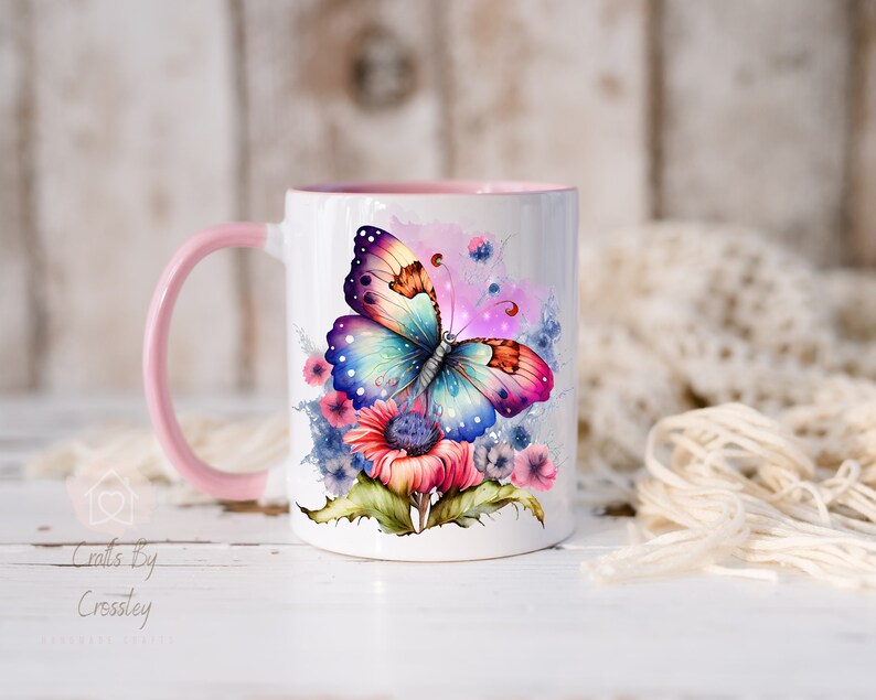 K&ouml;nnte beinhalten: Eine wei&szlig;e Keramiktasse mit einem rosa Henkel zeigt einen bunten Schmetterling mit schillernden Fl&uuml;geln und einem Blumenmuster. Der Schmetterling ist von rosa, blauen und gelben Blumen umgeben.