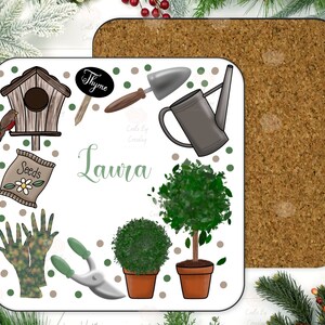 May include: A white square coaster with a gardening theme. It features a birdhouse, gardening gloves, a watering can, a pair of pruning shears, a bag of seeds, a small potted tree, a small potted bush, and a sign that says "Thyme". The name "Laura" is written in green cursive lettering.