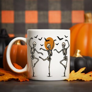 Pu&ograve; includere: Tazza in ceramica bianca con un design di Halloween con tre scheletri danzanti, pipistrelli e una luna piena. La tazza &egrave; ambientata su uno sfondo di zucche e foglie autunnali.