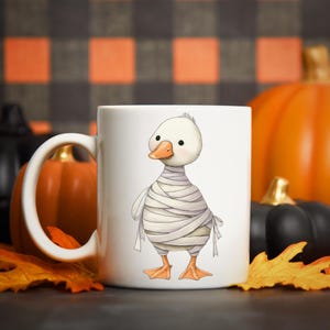 May include: White ceramic mug featuring a cartoon duck wrapped in bandages, resembling a mummy. The duck has an orange beak and feet. The mug is decorated with a Halloween theme, with pumpkins and fall leaves in the background.