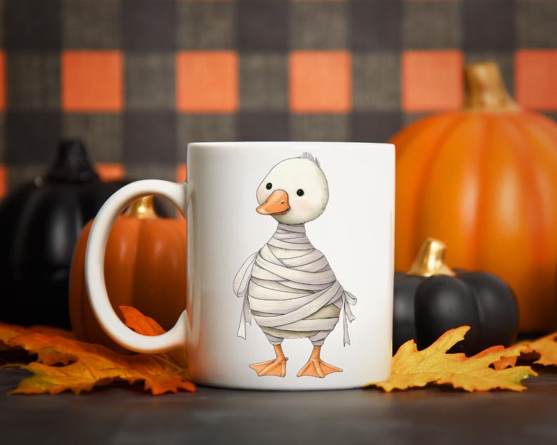 May include: White ceramic mug featuring a cartoon duck wrapped in bandages, resembling a mummy. The duck has an orange beak and feet. The mug is decorated with a Halloween theme, with pumpkins and fall leaves in the background.
